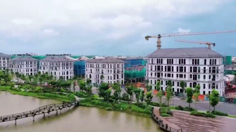 Dãy biệt thự Alluvia City đang xây bên hồ, có cầu gỗ đi bộ và cây xanh, chụp từ trên cao
