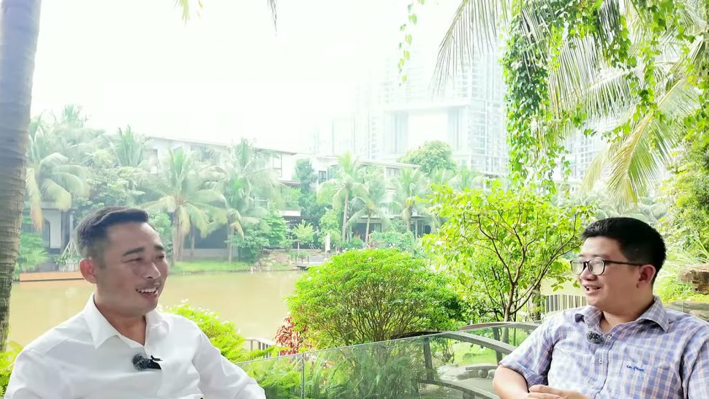 Hai người đàn ông ngồi trao đổi bên bàn kính cạnh bờ sông trong khu biệt thự đảo Ecopark, phía sau là dãy biệt thự thấp tầng và toà căn hộ cao tầng
