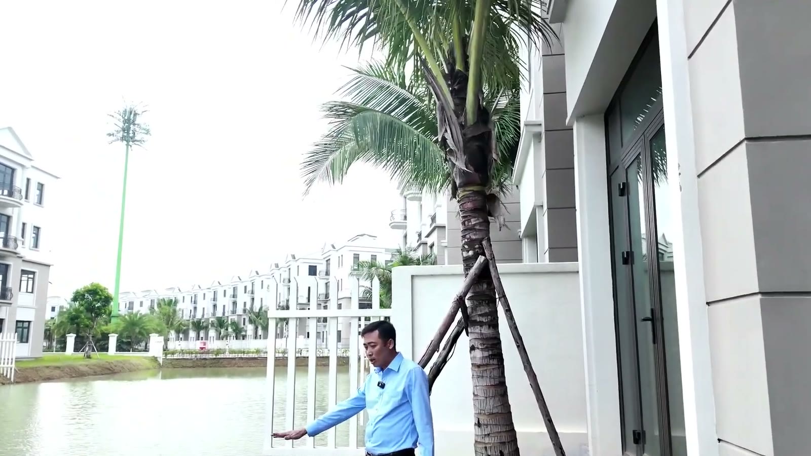 Lê Ngọc Lương đứng bên cây dừa được tặng ở phần đất sau một căn song lập Đảo Ngọc, phía trước là mặt nước và dãy nhà bên kia bờ