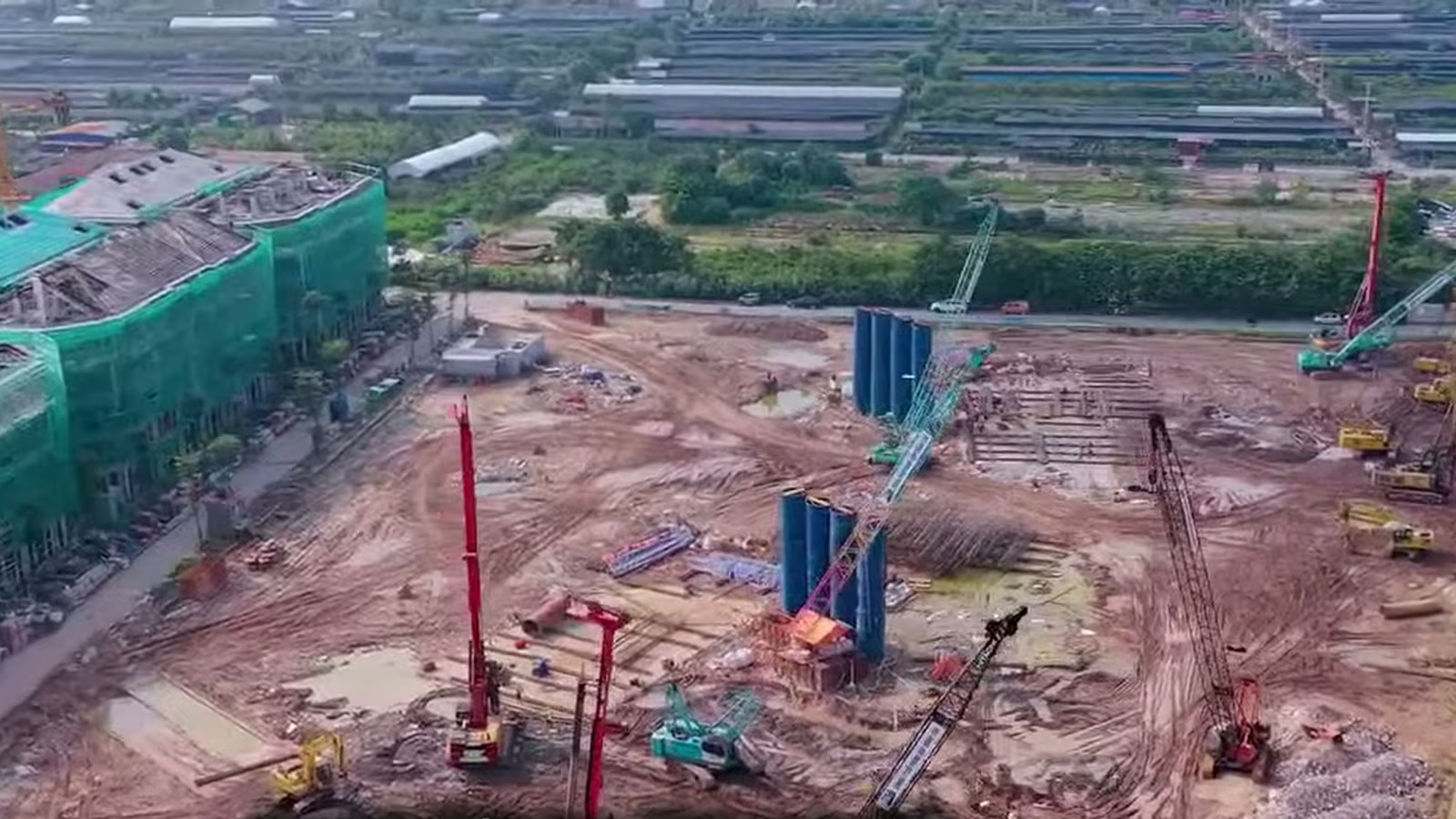 Công trường phần móng ở Alluvia City với nhiều máy ép cọc và cần cẩu đỏ đang làm việc trên nền đất đỏ, phía sau là khối nhà quây lưới xanh