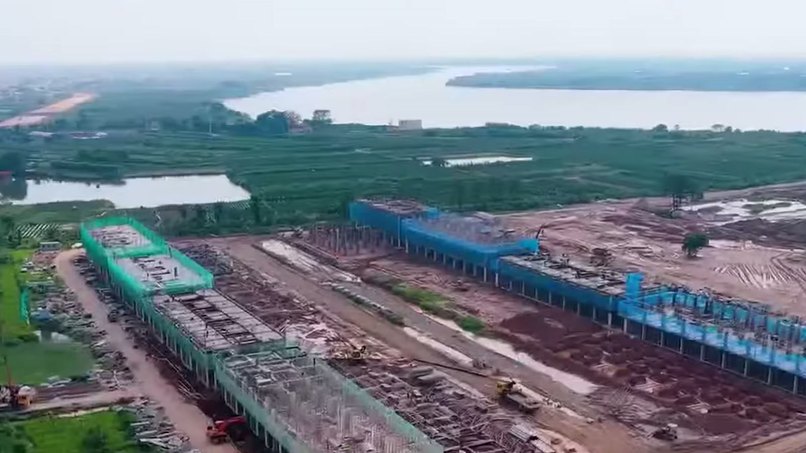 Khu vực ven sông trong Alluvia City đang thi công, hàng cọc và kết cấu bê tông chạy dọc mép nước, phía xa là sông Hồng và vùng cây xanh