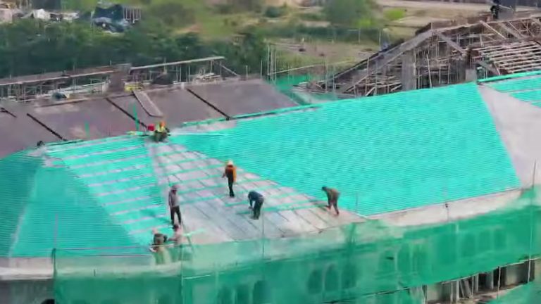 Thợ đang làm việc trên mái một dãy nhà thấp tầng Alluvia City, phần mái đã đổ và phủ lưới an toàn màu xanh, tháng 10/2026